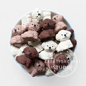 以下が含まれることがあります： 白、ベージュ、ダークブラウンの色合いをした、小さな3次元のプードル型チャームのコレクション。各チャームは、質感のあるカールした毛並み、黒い鼻と黒い目を持つ滑らかな顔が特徴です。チャームはボウルの中に入っています。