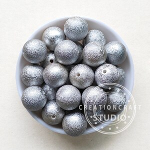 May include: A bowl filled with silver glitter-covered wooden beads. The beads are round and have a textured surface.