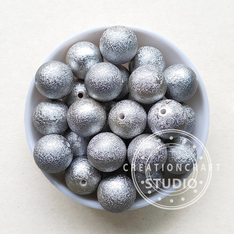 May include: A bowl filled with silver glitter-covered wooden beads. The beads are round and have a textured surface.