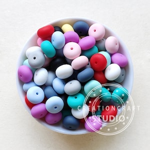 Pode incluir: Uma tigela cheia de contas de silicone coloridas em vários tons de azul, rosa, vermelho e cinza. As contas são lisas e têm uma forma ligeiramente arredondada.