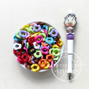 May include: A white bowl filled with colorful, wavy, donut-shaped polymer clay beads. The beads are in various colors, including red, orange, yellow, green, blue, purple, pink, brown, and white. A white pen with a purple and black Halloween-themed charm is next to the bowl.