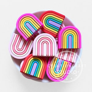 Pode incluir: Uma coleção de berloques de silicone em forma de arco-íris em uma tigela branca. Os berloques apresentam várias combinações de cores, incluindo listras rosa brilhante, amarelo, azul, roxo e vermelho. Uma marca d'água CREATIONCRAFT STUDIO é visível.