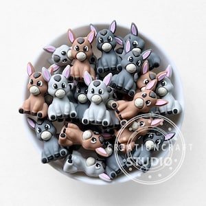 May include: A white bowl filled with small donkey figurines in various shades of gray and brown. The donkeys have pink ears and are in different poses. The Creation Craft Studio logo is visible on the bottom right.