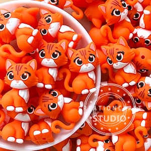 May include: A close-up of numerous orange cat-shaped embellishments with white details. Each cat has a smiling face, black eyes, and striped markings. Some are in a white bowl, with a logo that says "Creation Craft Studio".
