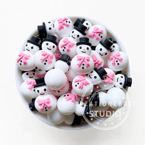 Pode incluir: Uma tigela cheia de enfeites brancos em forma de boneco de neve. Cada boneco de neve tem um chapéu preto, um laço rosa e olhos e boca de pontos pretos. Para artesanato e decoração de Natal.