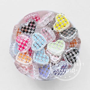 May include: A collection of heart-shaped beads in a white bowl. The beads are clear with woven patterns in various colors, including pink, blue, yellow, orange, and black. Some beads have a checkered design.