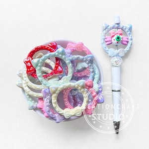 May include: A collection of colorful, cat-shaped rings in a white bowl, alongside a white pen with a cat-themed top. The rings are in shades of red, pink, blue, and purple. The pen features a decorative top with a bow and an eyeball design.