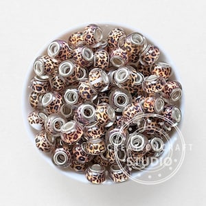 May include: A collection of leopard print glass beads in a white bowl. The beads have a clear center and silver-colored metal accents. The leopard print pattern is brown and black.