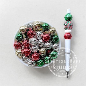 Puede incluir: Un tazón blanco lleno de cascabeles de diferentes colores: rojo, verde, dorado y plateado. Los cascabeles son de diferentes tamaños y formas. El tazón está sobre una superficie blanca. El texto "CREATIONCRAFT STUDIO" es visible en la esquina inferior derecha de la imagen.