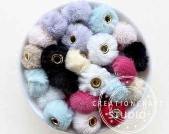 May include: A collection of colorful, fluffy pom-poms with gold-colored eyelets, displayed in a white bowl. The pom-poms are in various pastel shades, including pink, blue, lavender, and cream, with a few black and red ones.