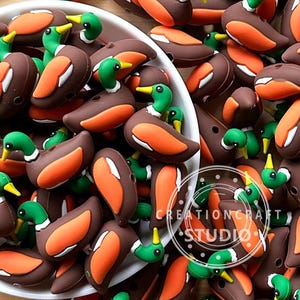 Könnte beinhalten: Nahaufnahme von zahlreichen Enten-förmigen Verzierungen in Braun-, Orange-, Grün- und Weißtönen. Die Enten haben braune Körper, orangefarbene Flügel, grüne Köpfe und gelbe Schnäbel. Die Verzierungen befinden sich in einer weißen Schüssel. Der Text "CREATIONCRAFT STUDIO" ist sichtbar.