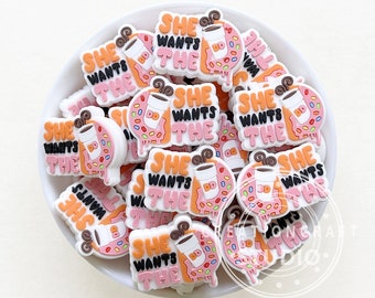 Könnte beinhalten: Eine Sammlung von weißen, rosa und orangefarbenen Donut-förmigen Charms mit dem Text "She Wants The" und einer Kaffeetassengrafik.