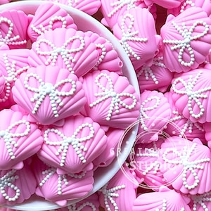 Peut inclure: Gros plan sur des embellissements roses en forme de coquillage avec des détails de nœuds blancs. Les coquillages sont dans un bol blanc et éparpillés autour. Les embellissements ont une surface texturée, probablement en résine ou similaire.