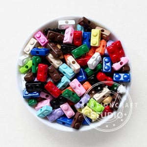 May include: A white bowl filled with a variety of colorful plastic building blocks. The blocks are in shades of red, blue, green, pink, brown, black, white, and yellow. The blocks are small and rectangular with rounded edges.