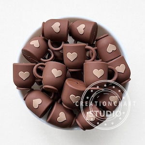 May include: A collection of small, brown mugs, each decorated with a light brown heart. The mugs are arranged in a white bowl. The text "CREATIONCRAFT STUDIO" is visible on the bottom right.