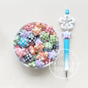 May include: A white bowl filled with colorful star-shaped beads. The beads have a checkered pattern in various colors, including pink, purple, green, orange, and black. The bowl is sitting on a white surface.