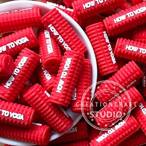 Puede incluir: Una colección de pequeños rodillos de masaje cilíndricos rojos con una superficie texturizada. Cada rodillo tiene el texto blanco "HOW TO YOGA" impreso. Los rodillos están dispersos en un cuenco blanco y alrededor.