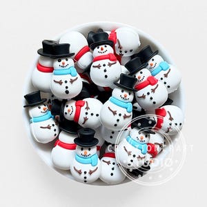Peut inclure: Une collection de petites perles blanches en forme de bonhomme de neige dans un bol blanc. Chaque bonhomme de neige porte un haut-de-forme noir, un nez en carotte orange, des bras en brindilles marron et des boutons noirs, avec des écharpes rouges ou bleu clair.