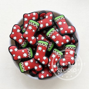 Puede incluir: Un bol lleno de abalorios de calcetines de Navidad rojos y verdes. Los calcetines tienen un patrón de copo de nieve blanco y un ribete de estampado de leopardo marrón y verde.