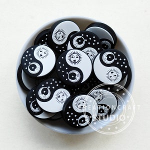 May include: A bowl filled with black and white silicone charms featuring a yin yang design with a sun and moon face on each side. The charms have a white background with black stars and a black background with white stars.
