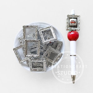 Puede incluir: Una colección de pequeños marcos de fotos plateados y ornamentados en un cuenco blanco. También se ve un bolígrafo blanco con una manzana roja y un pequeño dije de coche enmarcado. El bolígrafo tiene una punta plateada y un cuerpo blanco. Fondo blanco.