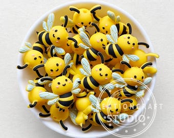May include: A collection of small, yellow and black bee-shaped charms with light blue wings and smiling faces, contained within a white bowl. The charms feature black antennae and stripes, with the text CREATIONCRAFT STUDIO visible in the lower right.