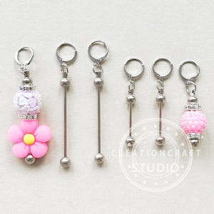 Könnte beinhalten: Fünf silberne Maschenmarkierer mit verschiedenen Anhängern. Die Anhänger umfassen eine rosa Blume, eine weiße und silberne Perle und eine rosa und silberne Perle. Die Maschenmarkierer sind an silbernen Ringen befestigt.