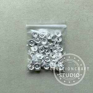 May include: A clear plastic bag filled with numerous silver-colored, faceted beads. The beads have a flower-like shape with a central hole. The bag is sealed and sits on a light gray surface. The image includes a watermark that says "CREATIONCRAFT STUDIO".