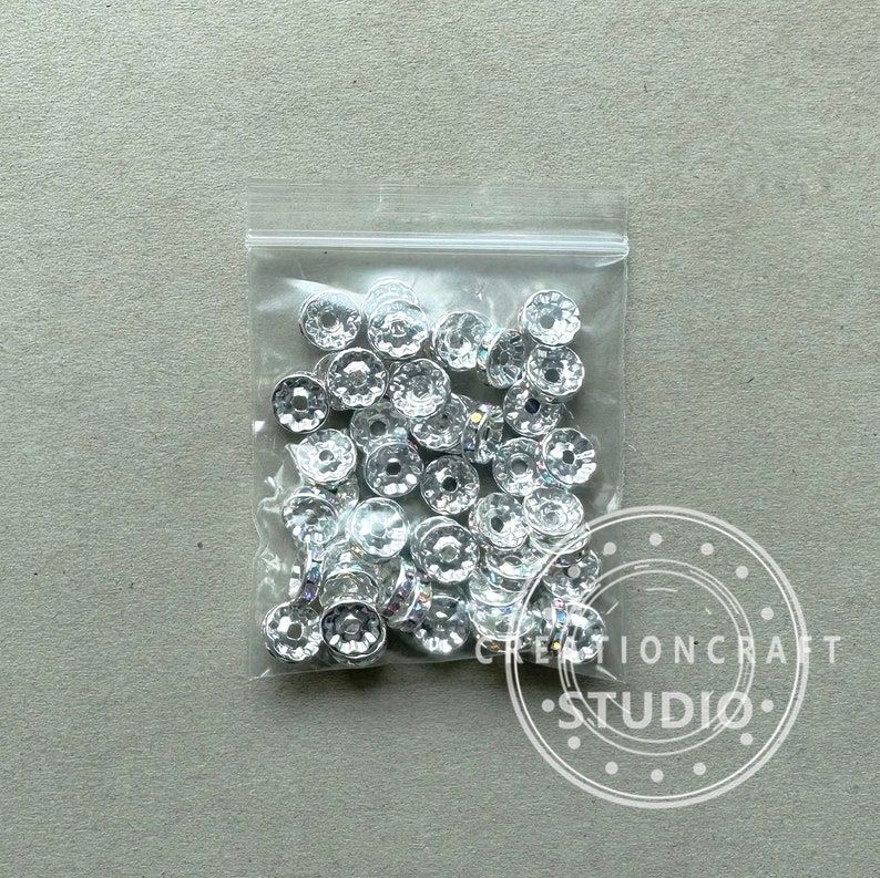 May include: A clear plastic bag filled with numerous silver-colored, faceted beads. The beads have a flower-like shape with a central hole. The bag is sealed and sits on a light gray surface. The image includes a watermark that says "CREATIONCRAFT STUDIO".