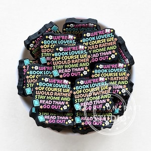 May include: A close-up of a pile of black silicone charms with the text "We're book lovers, of course we would rather stay home and read than go out." The charms are decorated with colourful flowers and books.
