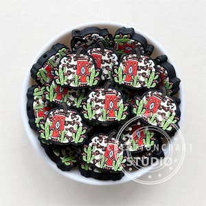 May include: A bowl filled with black and white silicone pumpkin charms. The pumpkins have a red and green geometric pattern and are surrounded by green cactus plants.