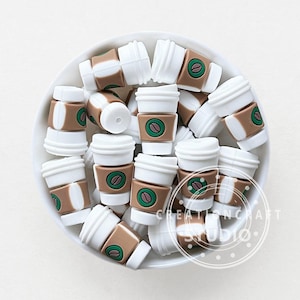 Puede incluir: Una colección de abalorios de taza de café en miniatura en un cuenco blanco. Los abalorios son blancos, marrones y verdes, con un diseño de grano de café. Los abalorios están dispuestos al azar, y el cuenco está sobre una superficie blanca.