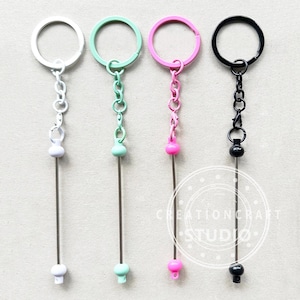 May include: Four keyrings in white, mint green, pink, and black. Each keyring has a matching coloured ring and chain, with a long, thin metal rod and a decorative bead at the bottom. These keyrings are for holding keys or small items.