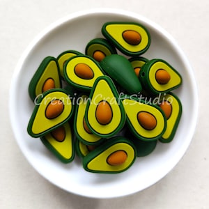 Puede incluir: Un cuenco blanco lleno de cuentas con forma de aguacate. Las cuentas son verdes, amarillas y marrones, con un contorno verde oscuro. El texto "CreationCraftStudio" es visible en el cuenco. Estas cuentas son ideales para proyectos de manualidades.