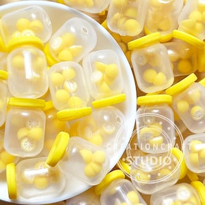 Pode incluir: Uma coleção de pequenos frascos de plástico transparente com tampas de rosca amarelo-brilhante. Cada frasco contém vários pingentes em miniatura em forma de fatias de limão amarelas. Os frascos estão dispostos numa tigela branca.