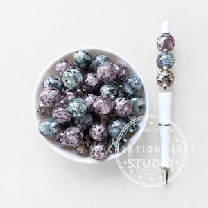 May include: A white bowl filled with clear, round beads with a floral design in shades of purple, blue, and silver. A white pen with matching decorative beads is also shown. The beads and pen are likely for crafting or jewelry making.