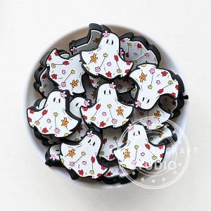 Op de afbeelding: Een kom gevuld met witte spookvormige bedels met bloemmotieven in roze, rood en gele tinten. De bedels zijn gemaakt van siliconen en zijn perfect om een spookachtige touch toe te voegen aan knutselwerk en projecten.