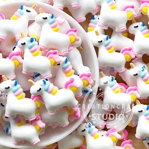 Op de afbeelding: Een kom vol witte unicorn-charms met regenboogaccenten. De charms zijn gemaakt van polymer clay en hebben een glanzende afwerking.