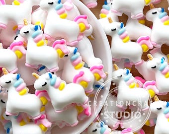 以下が含まれることがあります： 虹色のアクセントが付いた白いユニコーンチャームが詰まったボウル。チャームはポリマークレイ製で、光沢のある仕上げになっています。