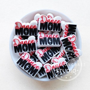 Peut inclure: Un bol rempli de bracelets à breloques blancs et noirs avec le texte "Dance Mom" en lettres rouges et noires.
