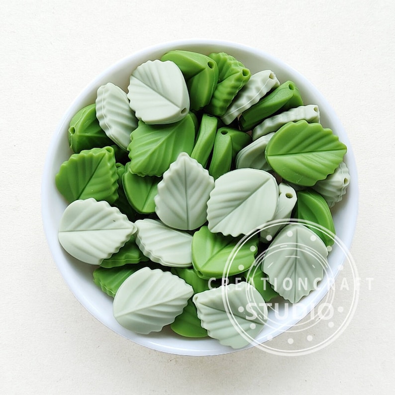 May include: A white bowl filled with green and light green leaf-shaped beads. The beads have detailed veining and are made of a smooth material. The beads are a mix of light and dark green shades. The bowl is on a white surface.