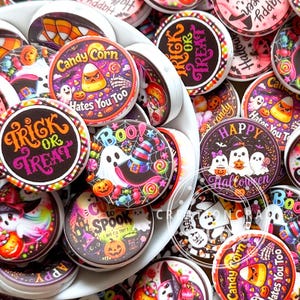 Può includere: Una collezione di bottoni rotondi e piatti a tema Halloween con grafiche colorate di fantasmi, zucche, caramelle a forma di mais e cappelli da strega. I disegni includono testi come Trick or Treat, Candy Corn Hates You Too, Boo e Happy Halloween.