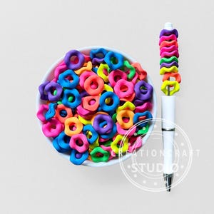Op de afbeelding: Een witte kom gevuld met kleurrijke, ringvormige polymeerklei kralen in roze, oranje, geel, groen, blauw en paars. Er is ook een witte pen met een regenboogkleurig polymeerklei ontwerp. Het ontwerp van de pen heeft gestapelde, ringvormige kralen.