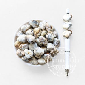 May include: A white ceramic bowl filled with small, beige and white seashells. A white pen with seashell accents is positioned to the right. The pen has a silver band and a silver tip. The seashells are a mix of colors and textures.