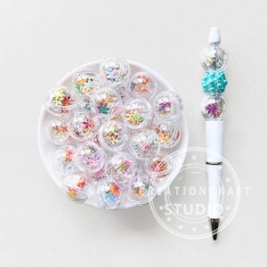 May include: A white pen with a decorative top featuring clear globes filled with colorful confetti and a turquoise bead. A white bowl holds numerous clear plastic globes filled with confetti.