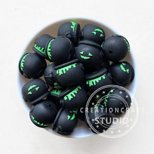 May include: A collection of black Halloween-themed beads in a white bowl. The beads are shaped like cauldrons and have green accents, including dripping liquid and jack-o'-lantern faces. The image includes the text "CREATIONCRAFT STUDIO".
