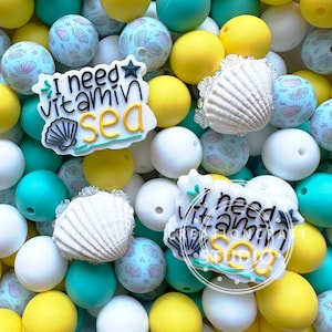 Pode incluir: Um close-up de duas contas de silicone coloridas com o texto "I need vitamin sea" nelas. As contas são cercadas por outras contas de silicone de várias cores, incluindo amarelo, branco e turquesa.