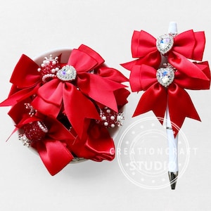 May include: A collection of red satin bows with heart-shaped rhinestone embellishments. Some bows are in a white bowl, while others adorn a white pen. The bows have multiple layers and are accented with small pearls and rhinestones. The pen has two bows attached.