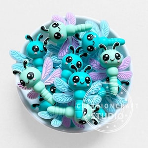 Può includere: Una collezione di piccoli ciondoli a forma di libellula color turchese con grandi occhi neri, guance rosa e ali viola chiaro. I ciondoli sono raccolti in una ciotola bianca ed è visibile un logo circolare con il testo CREATIONCRAFT STUDIO.