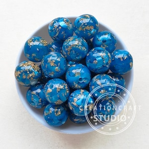 May include: A bowl of teal blue and gold beads. The beads are round and have a marbled effect with gold flecks throughout.